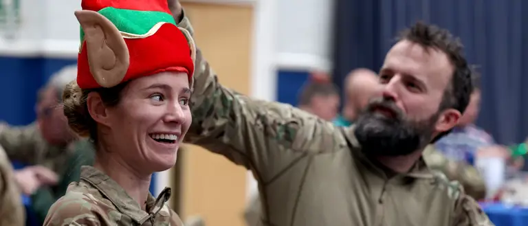 Two soldiers wearing camouflage uniforms sit next to each other. The one on the right fixes a Christmas hat of the one on the left.