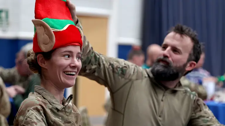 Two soldiers wearing camouflage uniforms sit next to each other. The one on the right fixes a Christmas hat of the one on the left.