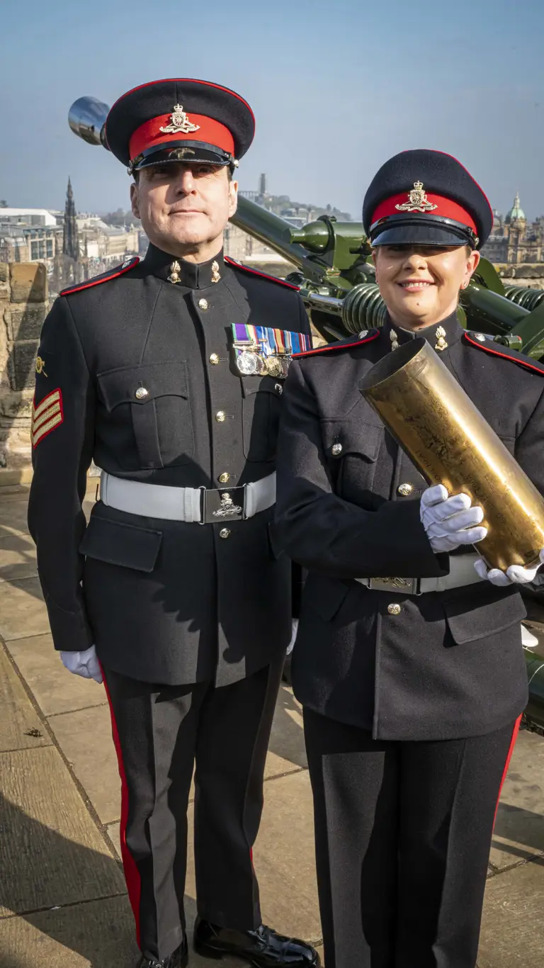 Gunner Caroline Gallagher is stood next to full time District Gunner Dave Beveridge at Edinburgh Castle, both dressed in No.1 uniform dress. Gallagher is holding an empty shell from the gun salute.