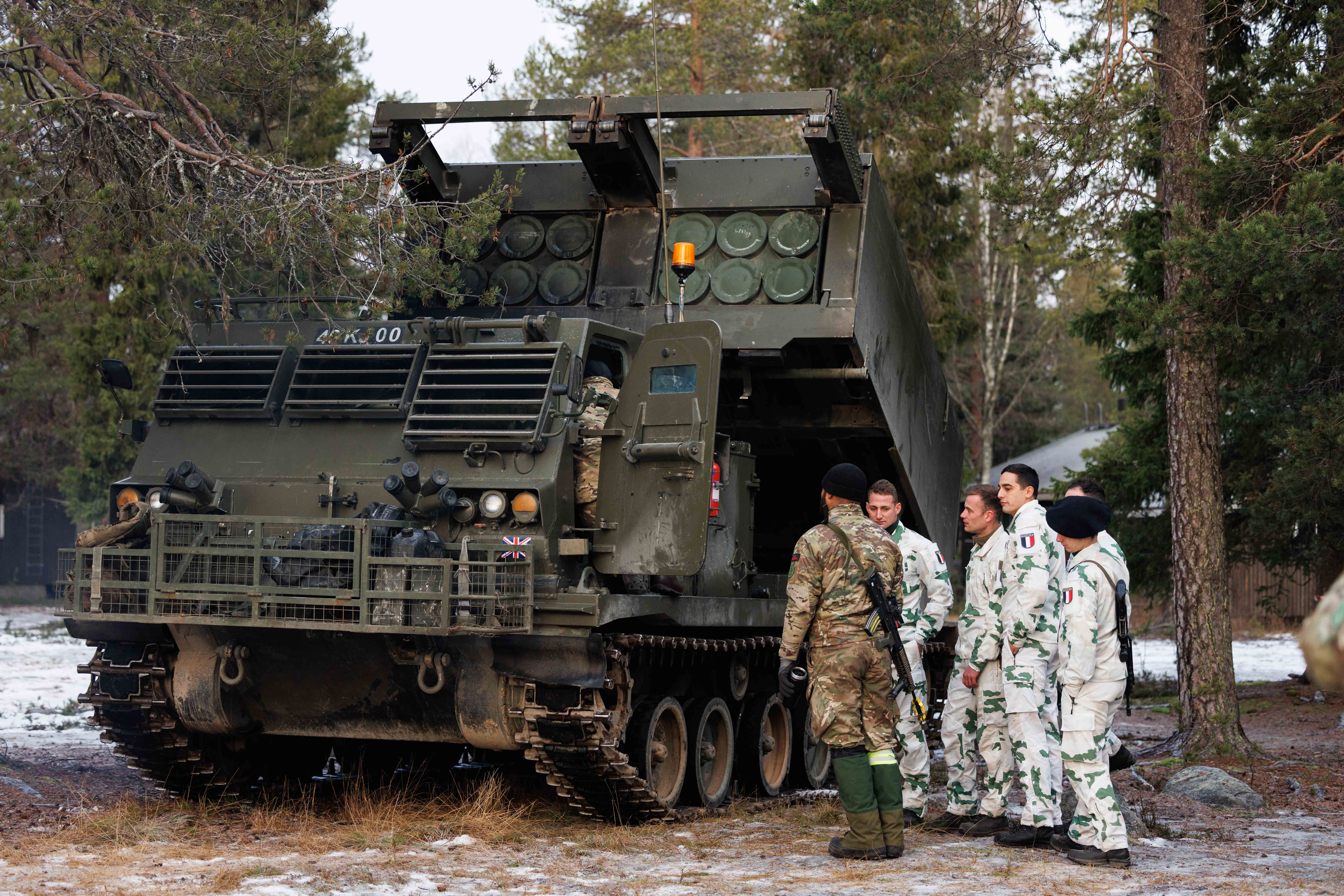 A British soldier in uniform is accompanied by several French soldiers in uniform, they are stood next to a vehicle that launches rockets.