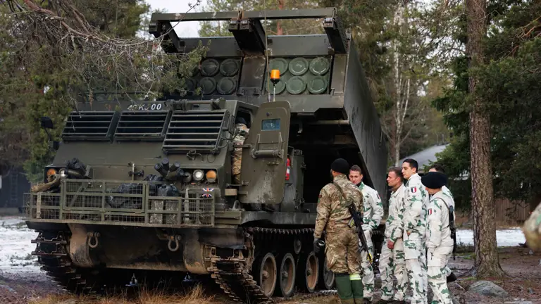 A British soldier in uniform is accompanied by several French soldiers in uniform, they are stood next to a vehicle that launches rockets.