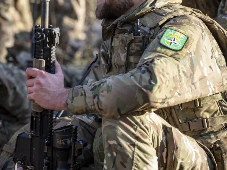Soldier in camouflage uniform shown sitting down for rest holding his rifle up. The soldier has a NATO Allied Reaction Force flash on his arm.