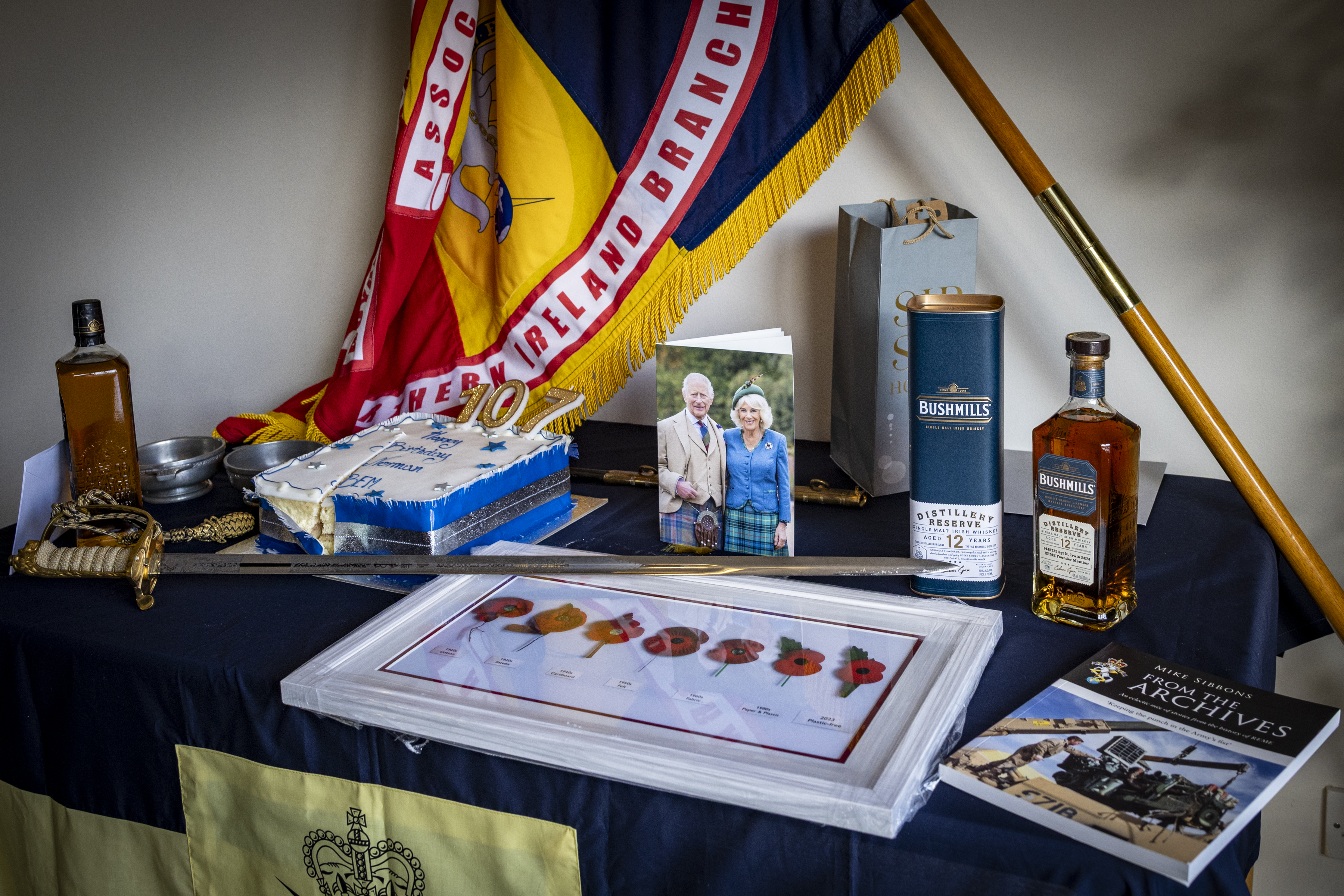 Memorial display featuring a flag, framed poppy artwork, bottles of Bushmills whiskey, and a photo of an elderly couple on a table.