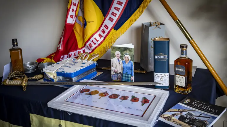 Memorial display featuring a flag, framed poppy artwork, bottles of Bushmills whiskey, and a photo of an elderly couple on a table.