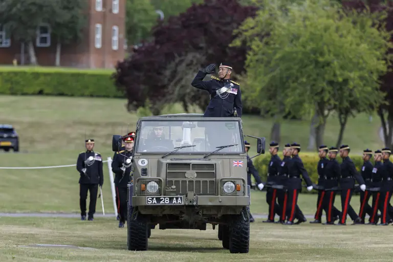 A person in a formal military uniform stands in an open-top vehicle, saluting. The vehicle has a British flag on its front. 