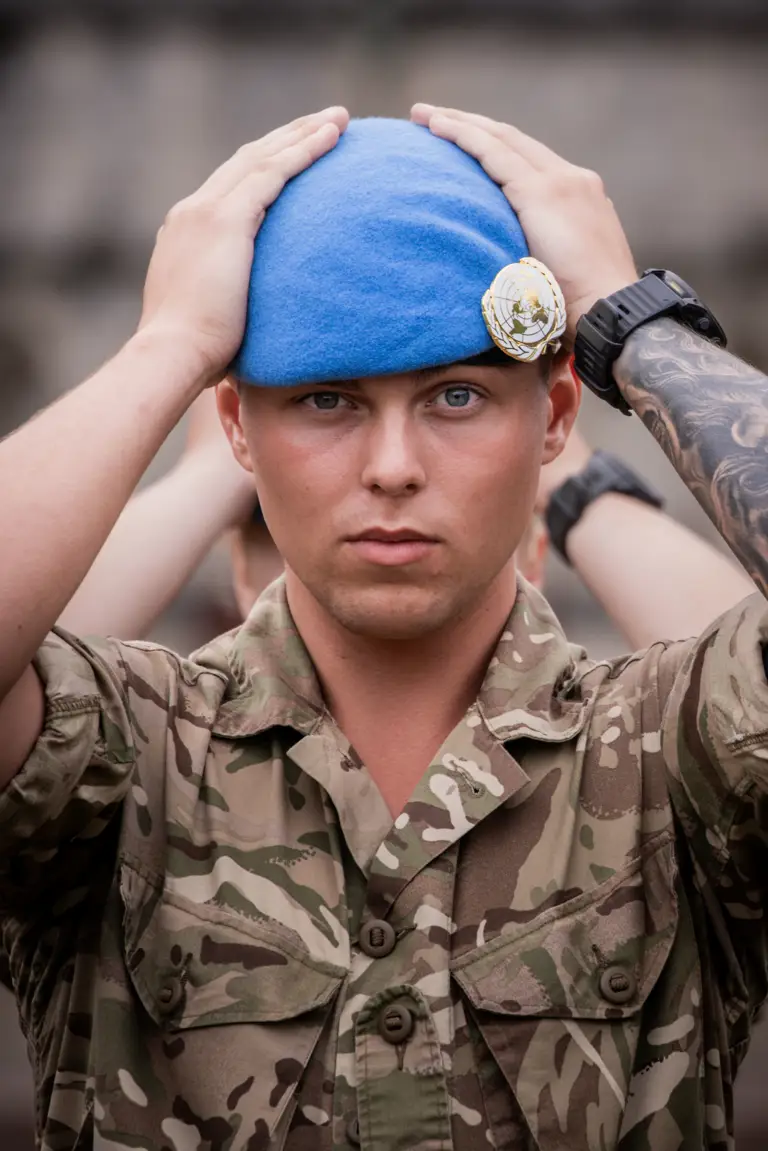 Soldier in camouflage uniform adjusting a blue beret on their head with both hands. 