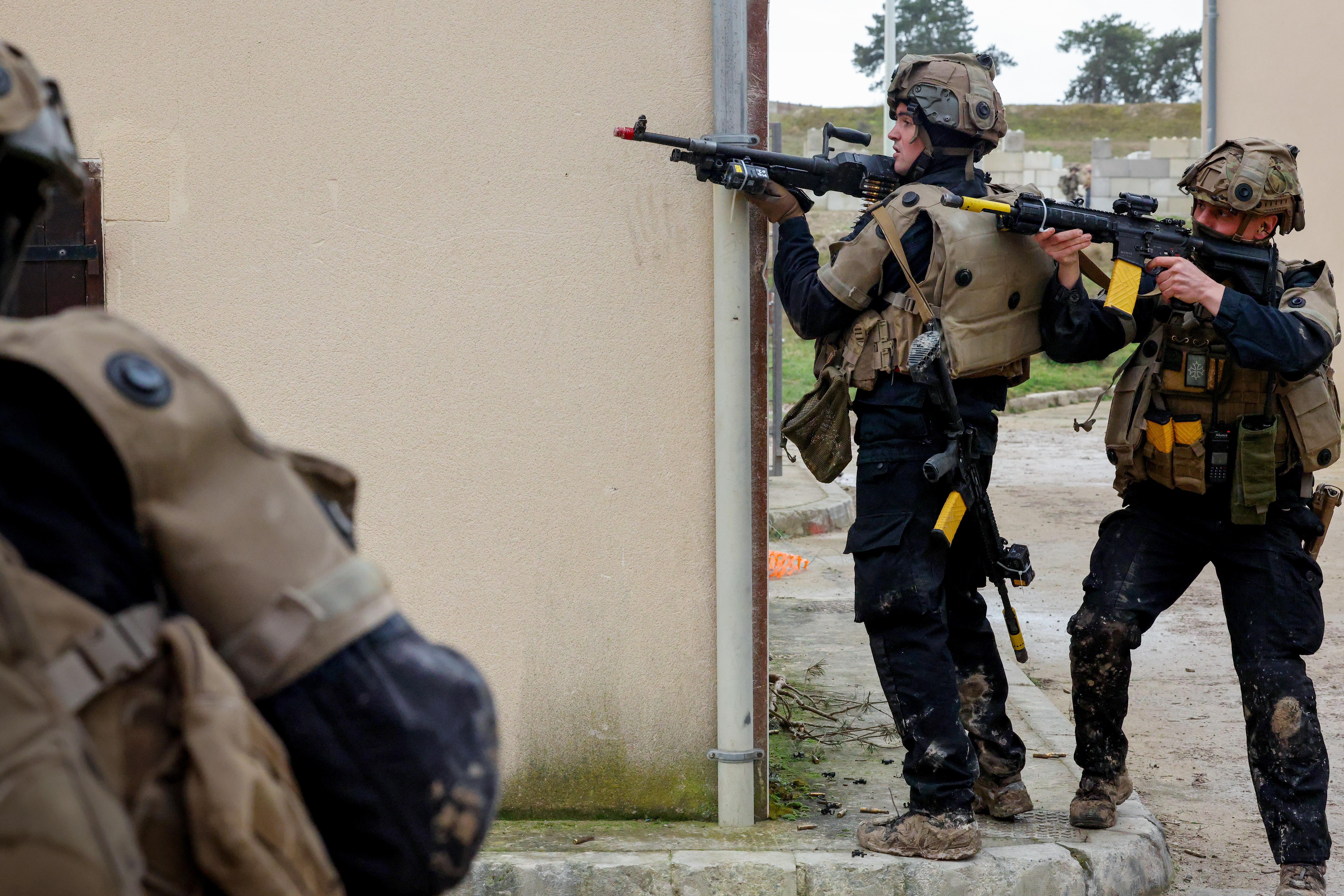 Two soldiers in tactical gear aiming rifles around a corner of a beige building during a training exercise.