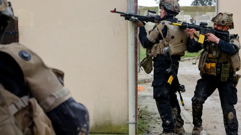Two soldiers in tactical gear aiming rifles around a corner of a beige building during a training exercise.