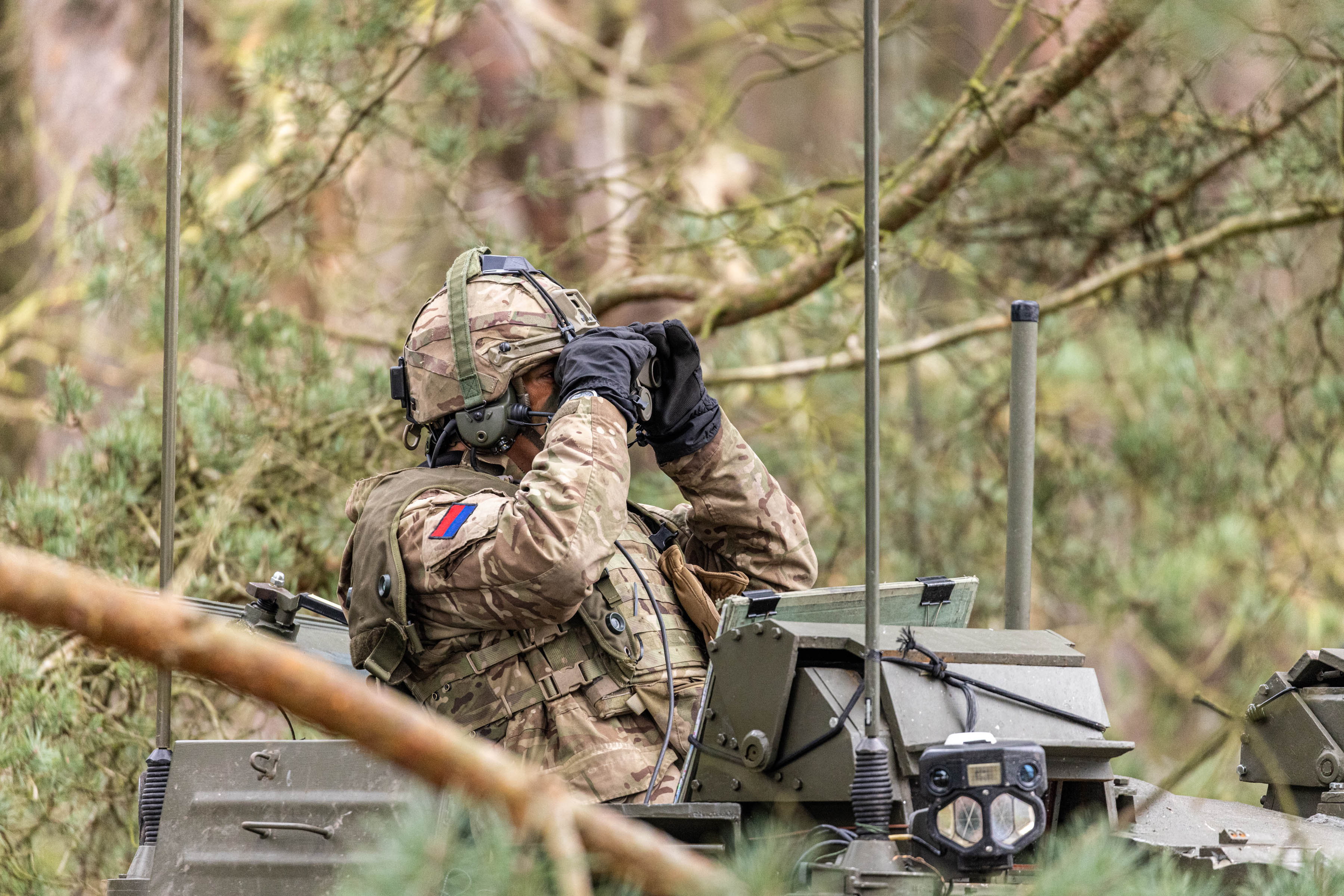 A soldier in camouflage uniform and helmet sits atop a military vehicle in a forested area, using binoculars. 