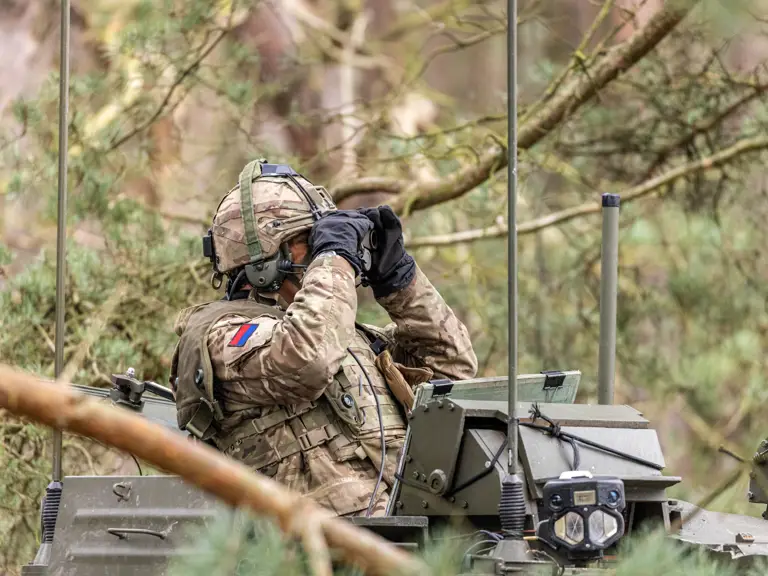 A soldier in camouflage uniform and helmet sits atop a military vehicle in a forested area, using binoculars.