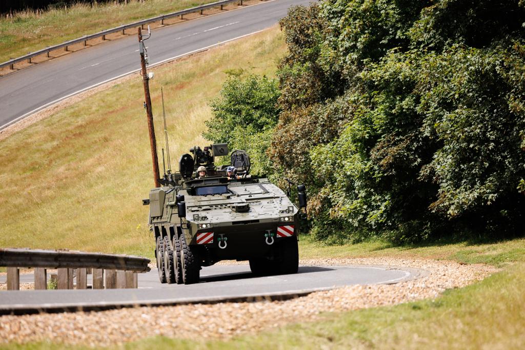 A military vehicle is driving along a curved paved road, surrounded by grassy areas and dense trees.