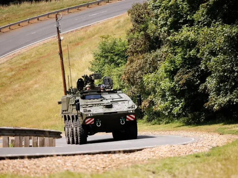 A military vehicle is driving along a curved paved road, surrounded by grassy areas and dense trees.