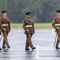 Four individuals in brown military uniforms and hats are marching in formation on a wet parade ground. They carry ceremonial canes and have red sashes. The background is a grassy area with trees.