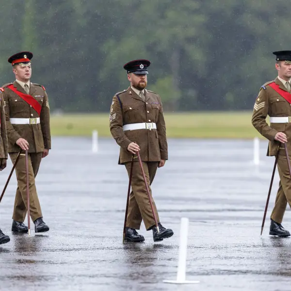 Four individuals in brown military uniforms and hats are marching in formation on a wet parade ground. They carry ceremonial canes and have red sashes. The background is a grassy area with trees.