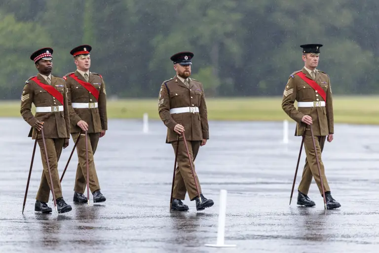Four individuals in brown military uniforms and hats are marching in formation on a wet parade ground. They carry ceremonial canes and have red sashes. The background is a grassy area with trees.