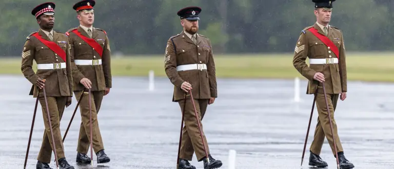Four individuals in brown military uniforms and hats are marching in formation on a wet parade ground. They carry ceremonial canes and have red sashes. The background is a grassy area with trees.