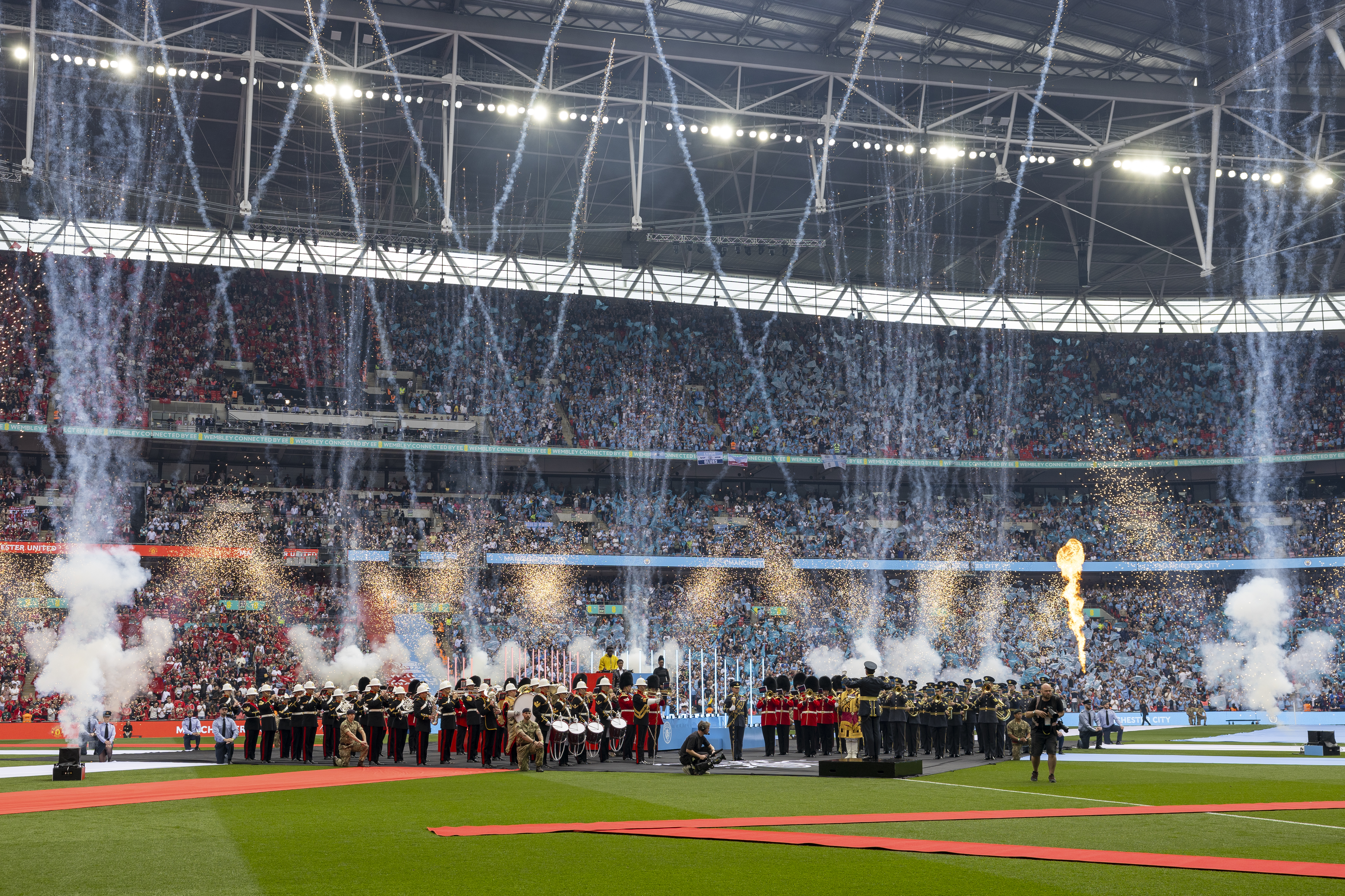 A large crowd fills Wembley Stadium, with a football field in the foreground. Fireworks and pyrotechnics explode. 