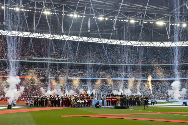 A large crowd fills Wembley Stadium, with a football field in the foreground. Fireworks and pyrotechnics explode.