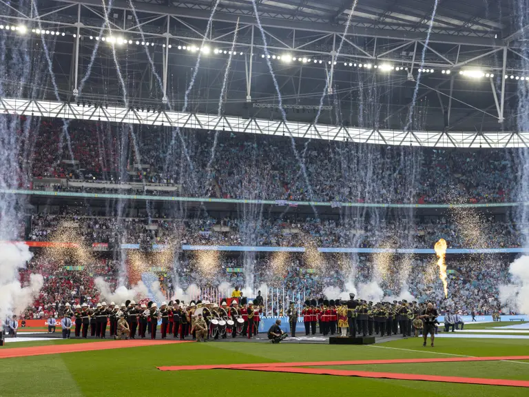 A large crowd fills Wembley Stadium, with a football field in the foreground. Fireworks and pyrotechnics explode.