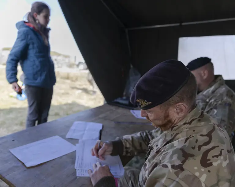 A military officer in camouflage writes at a table inside a tent, while a woman in a blue jacket stands outside, holding a small object. Calm, focused atmosphere.