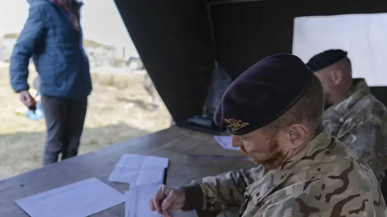 A military officer in camouflage writes at a table inside a tent, while a woman in a blue jacket stands outside, holding a small object. Calm, focused atmosphere.