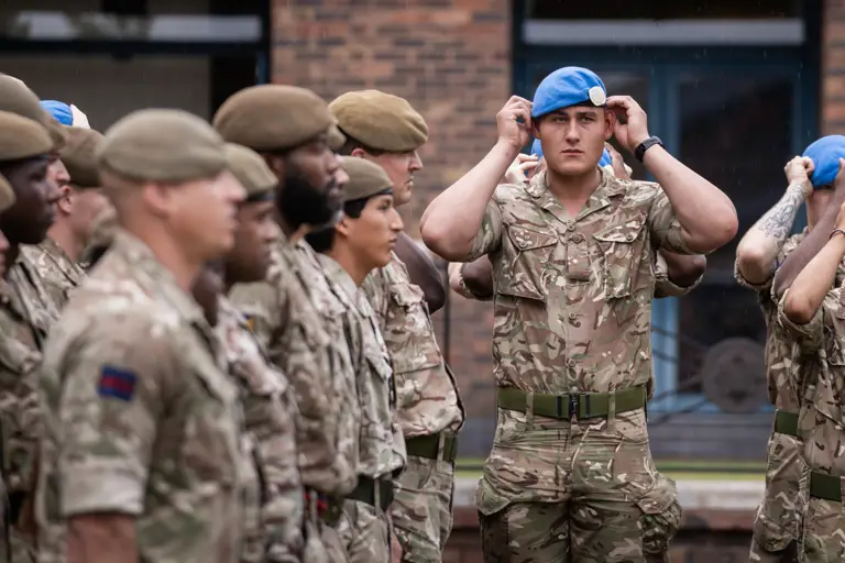 Soldier in camouflage uniform adjusting a blue beret on their head with both hands. Beside him soldiers stand wearing khaki berets on their heads.