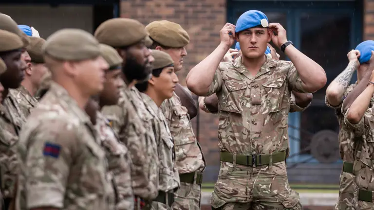 Soldier in camouflage uniform adjusting a blue beret on their head with both hands. Beside him soldiers stand wearing khaki berets on their heads.