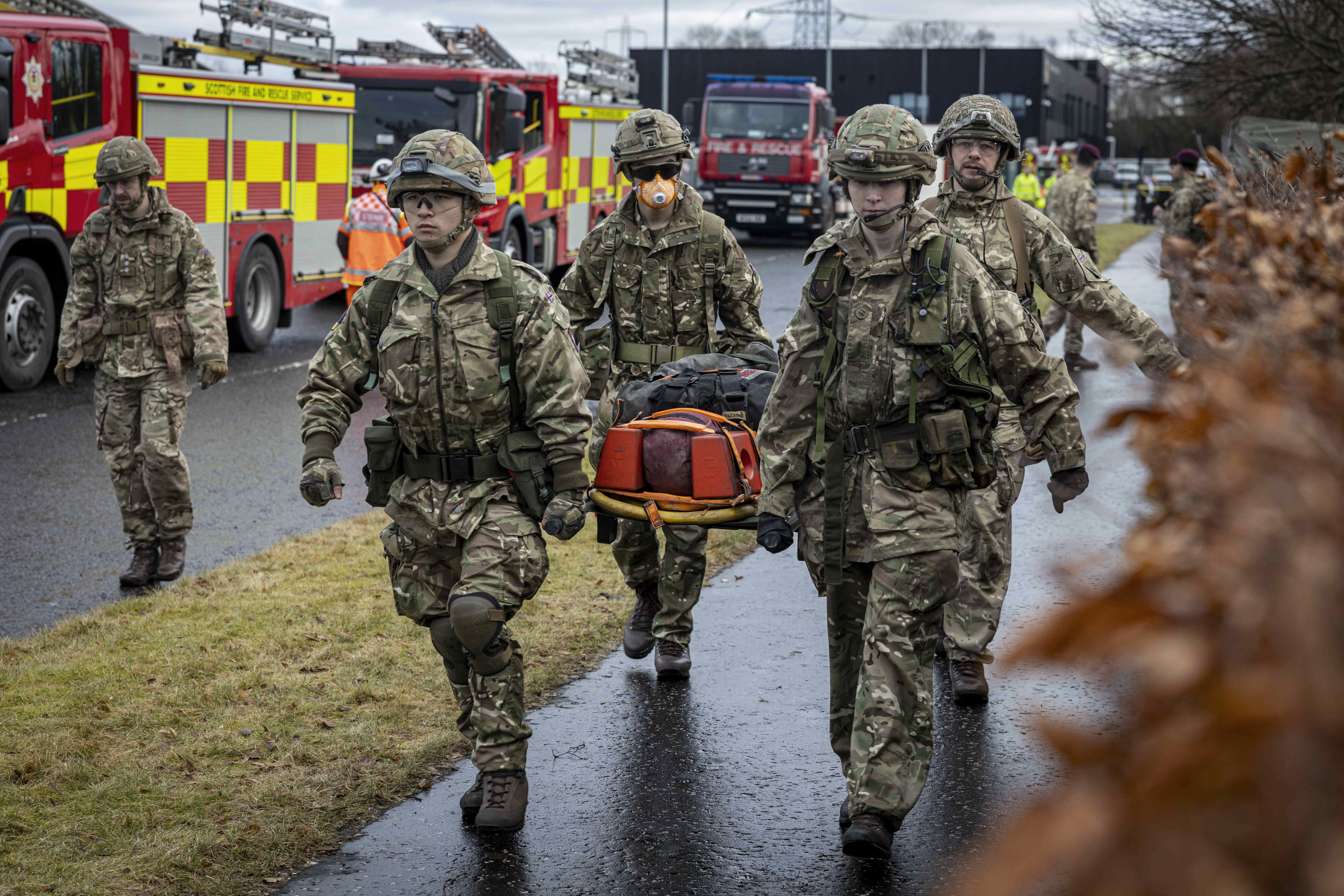 Soldiers in camouflage uniforms carry a stretcher with an injured person near fire engines on a wet road.