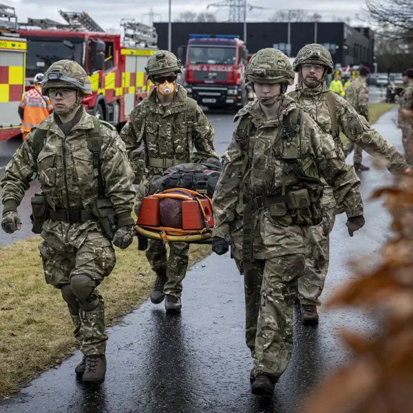 Soldiers in camouflage uniforms carry a stretcher with an injured person near fire engines on a wet road.