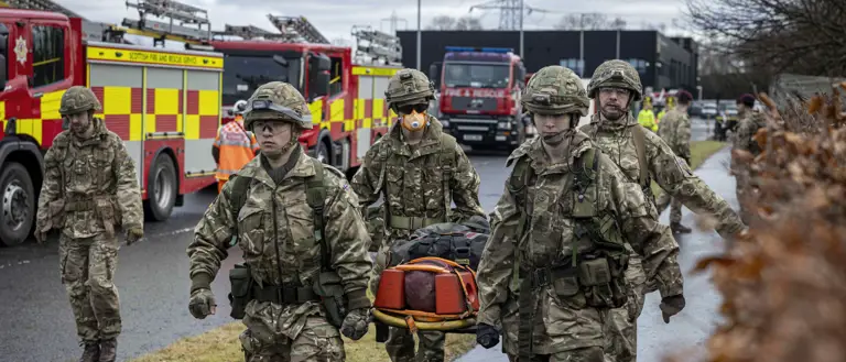 Soldiers in camouflage uniforms carry a stretcher with an injured person near fire engines on a wet road.