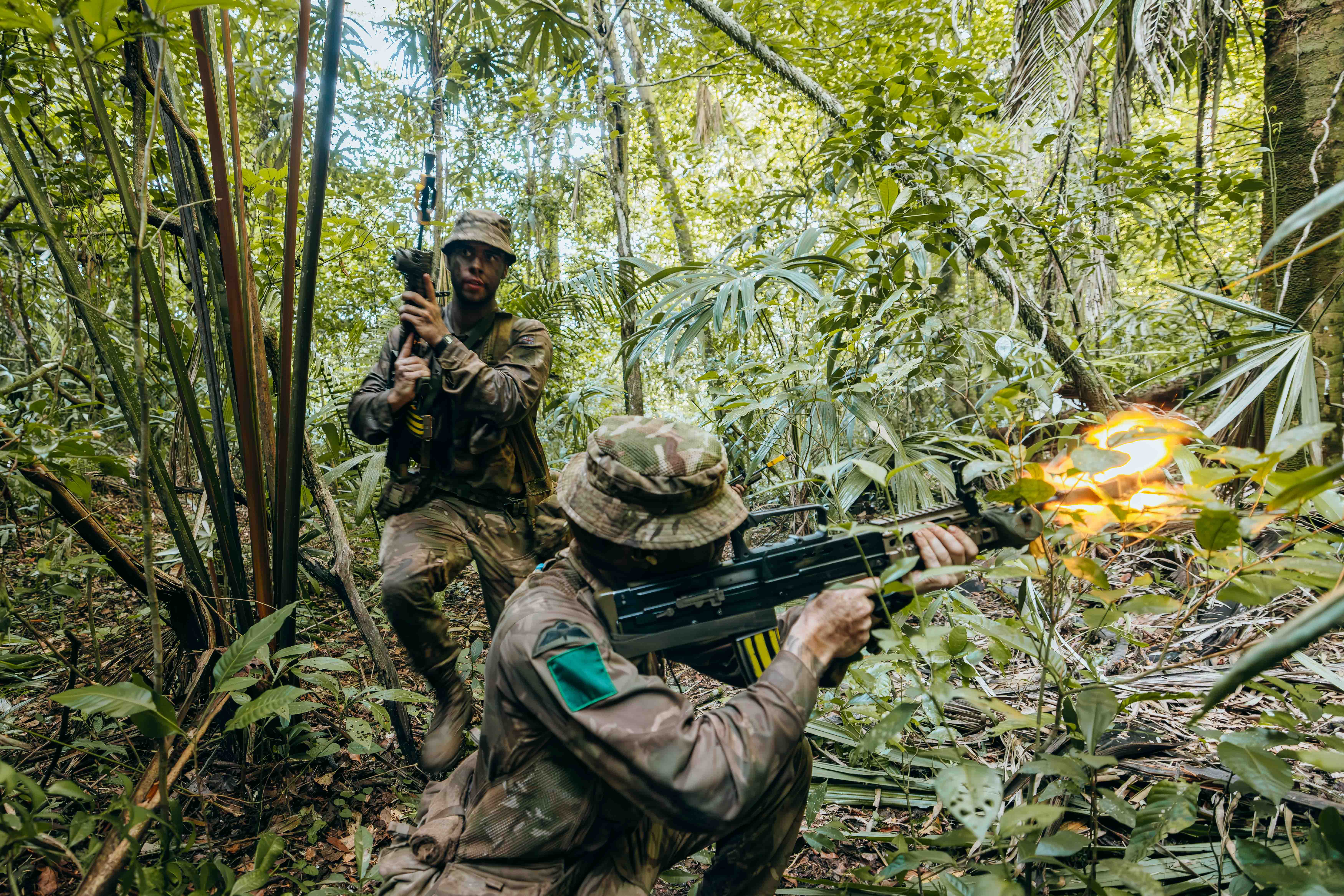 Two soldiers in camouflage gear firing weapons while navigating dense jungle foliage during a combat exercise.