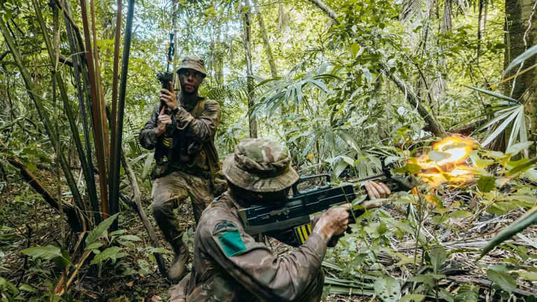 Two soldiers in camouflage gear firing weapons while navigating dense jungle foliage during a combat exercise.