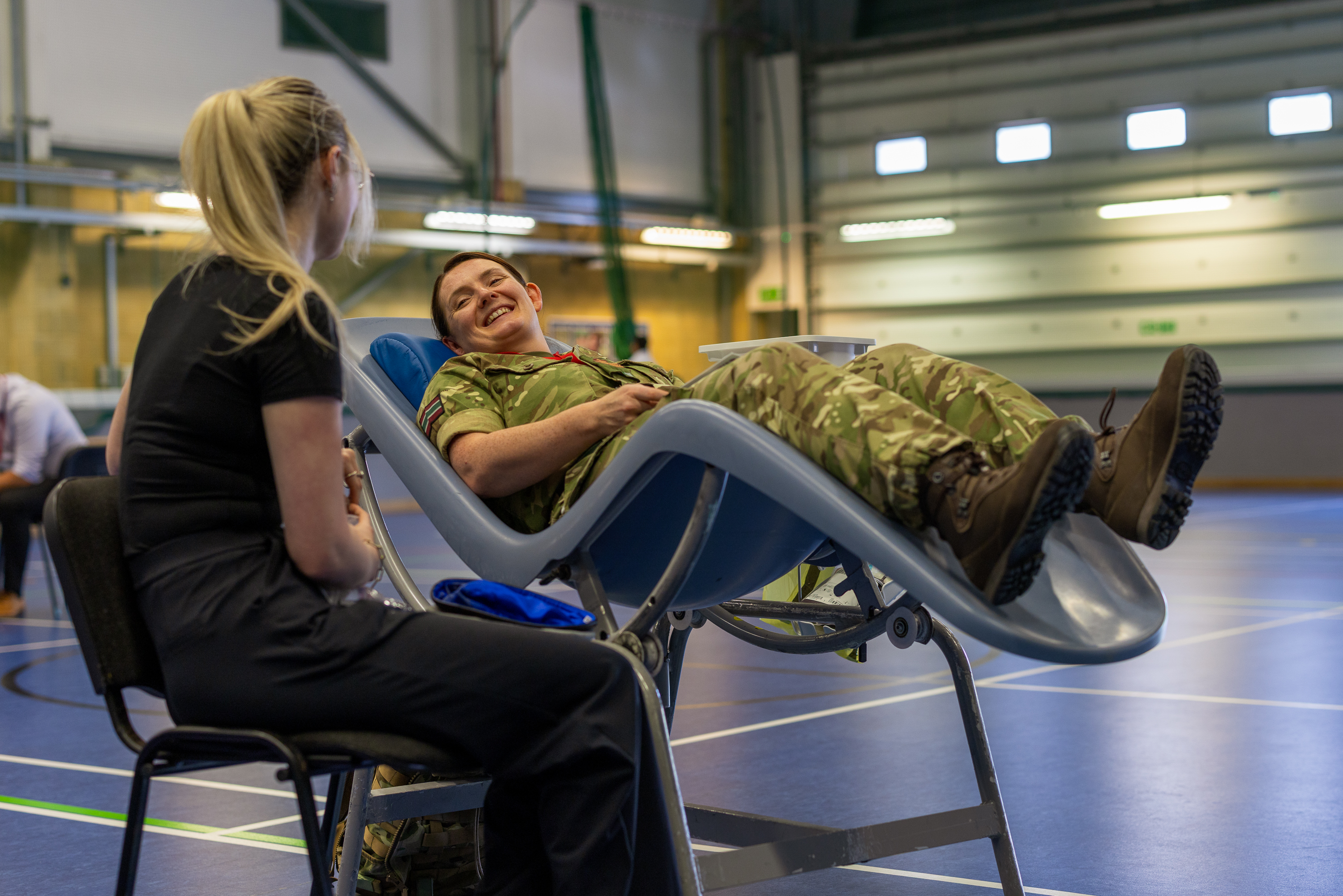 Soldier in camouflage uniform reclines on a medical chair inside a gymnasium while a woman in black sits nearby.