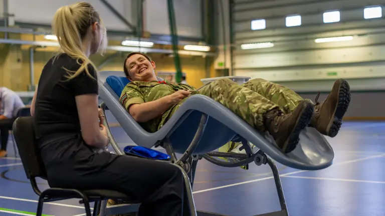 Soldier in camouflage uniform reclines on a medical chair inside a gymnasium while a woman in black sits nearby.