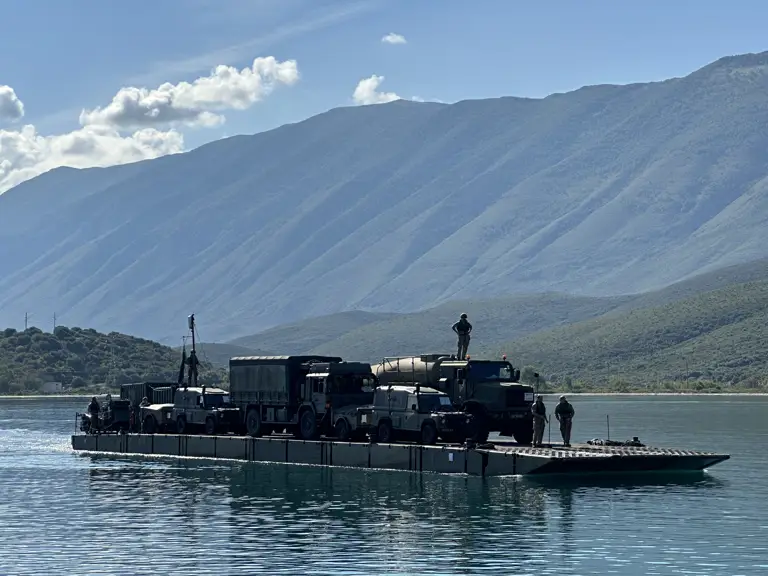 A soldier stands on top of a truck which sits on a ferry during a water crossing.