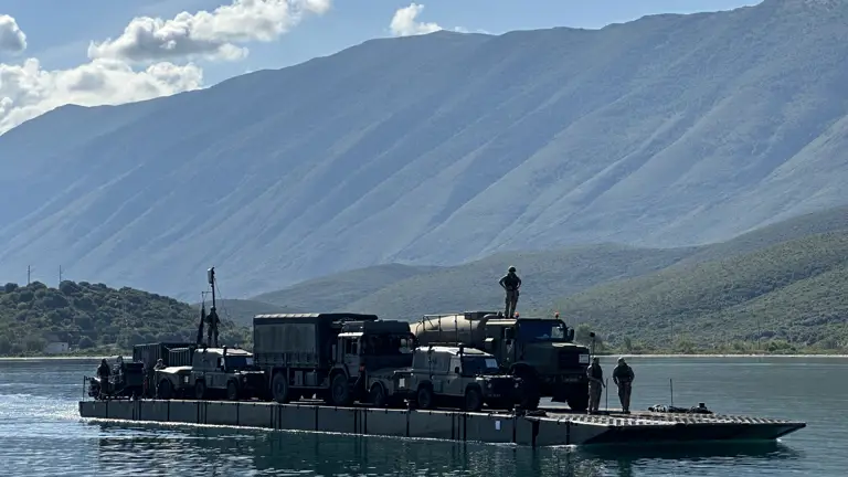 A soldier stands on top of a truck which sits on a ferry during a water crossing.