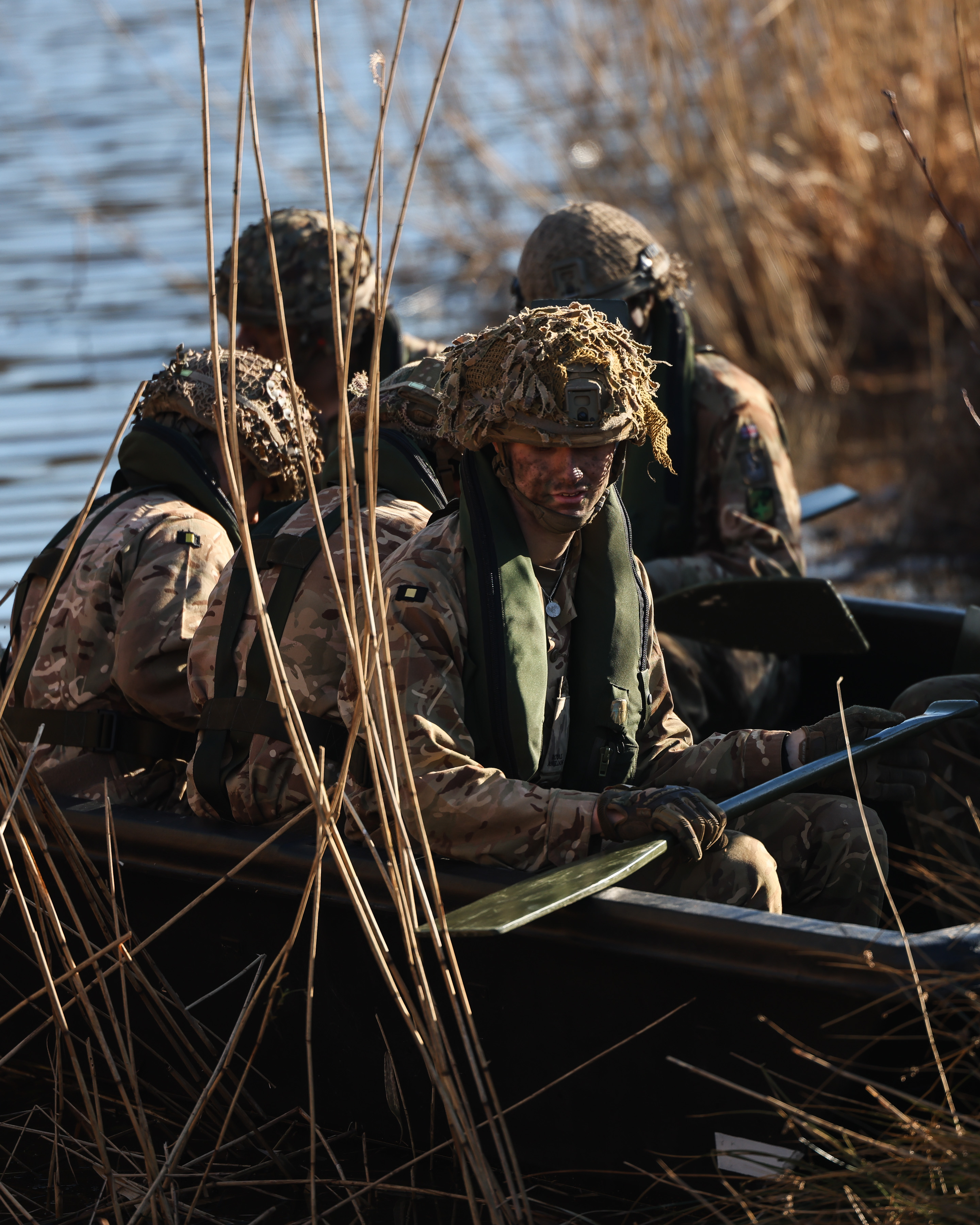 Five soldiers in full exercise kit and helmets are seen wearing a life jacket while on a small boat with paddles while attempting a river crossing