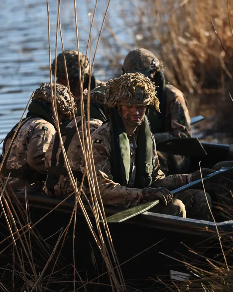 Five soldiers in full exercise kit and helmets are seen wearing a life jacket while on a small boat with paddles while attempting a river crossing
