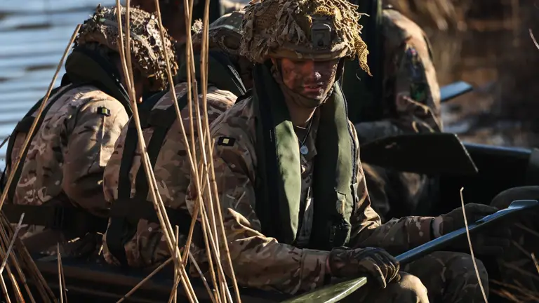 Five soldiers in full exercise kit and helmets are seen wearing a life jacket while on a small boat with paddles while attempting a river crossing