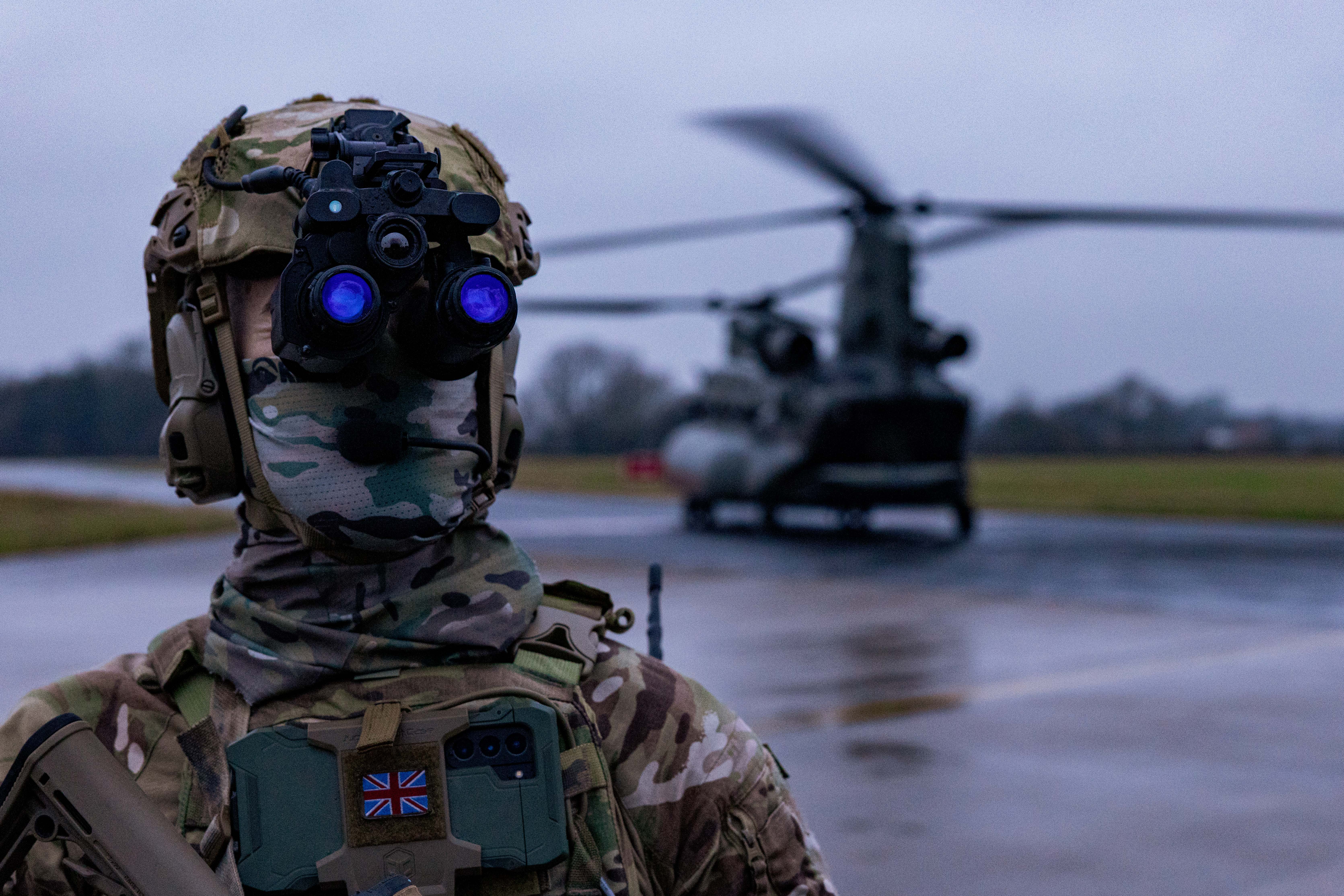Soldier in full camouflage gear with night vision goggles stands on wet tarmac near a military helicopter.