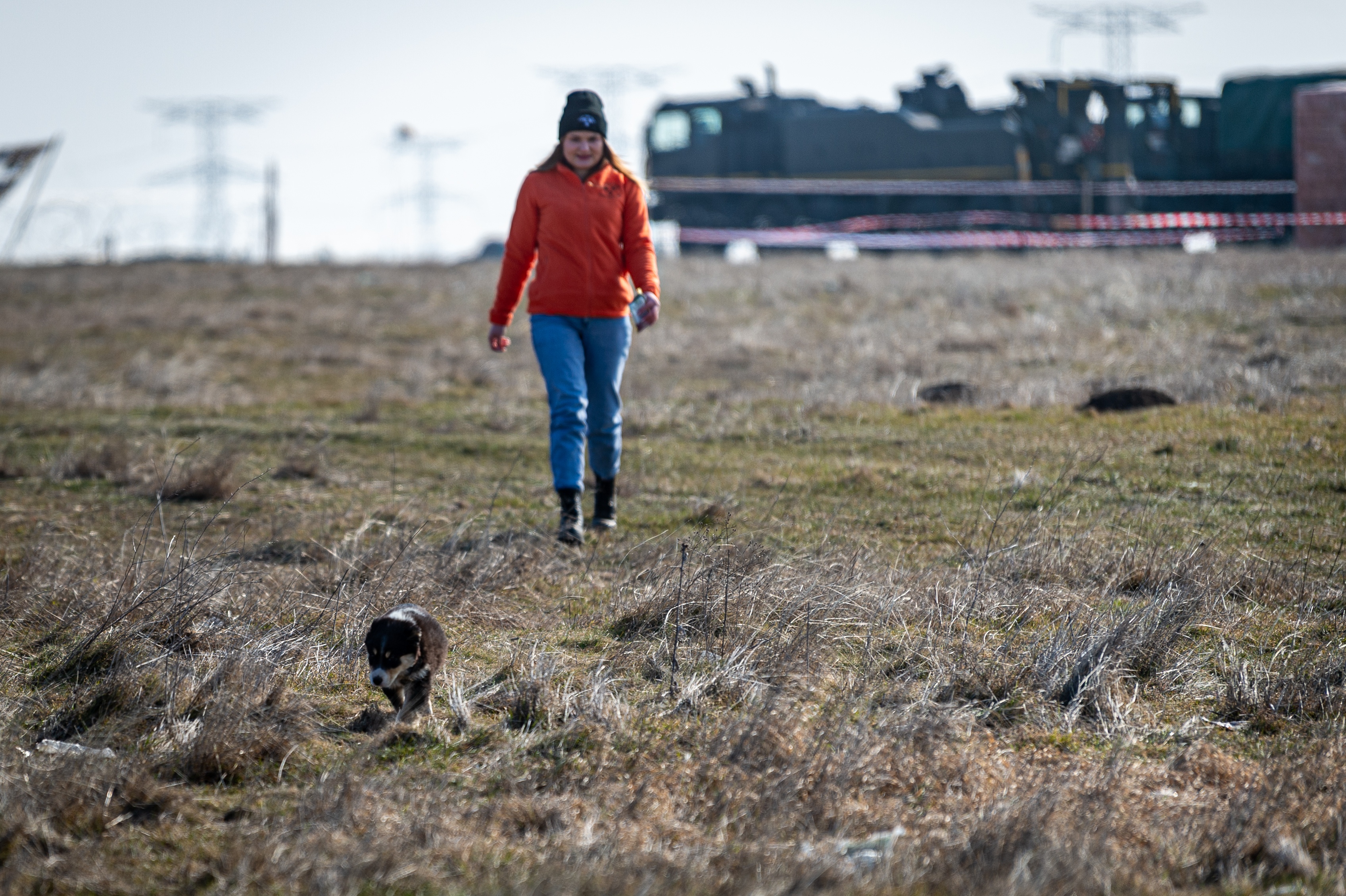 A lady in casual civilian attire walks behind a puppy in a field. 