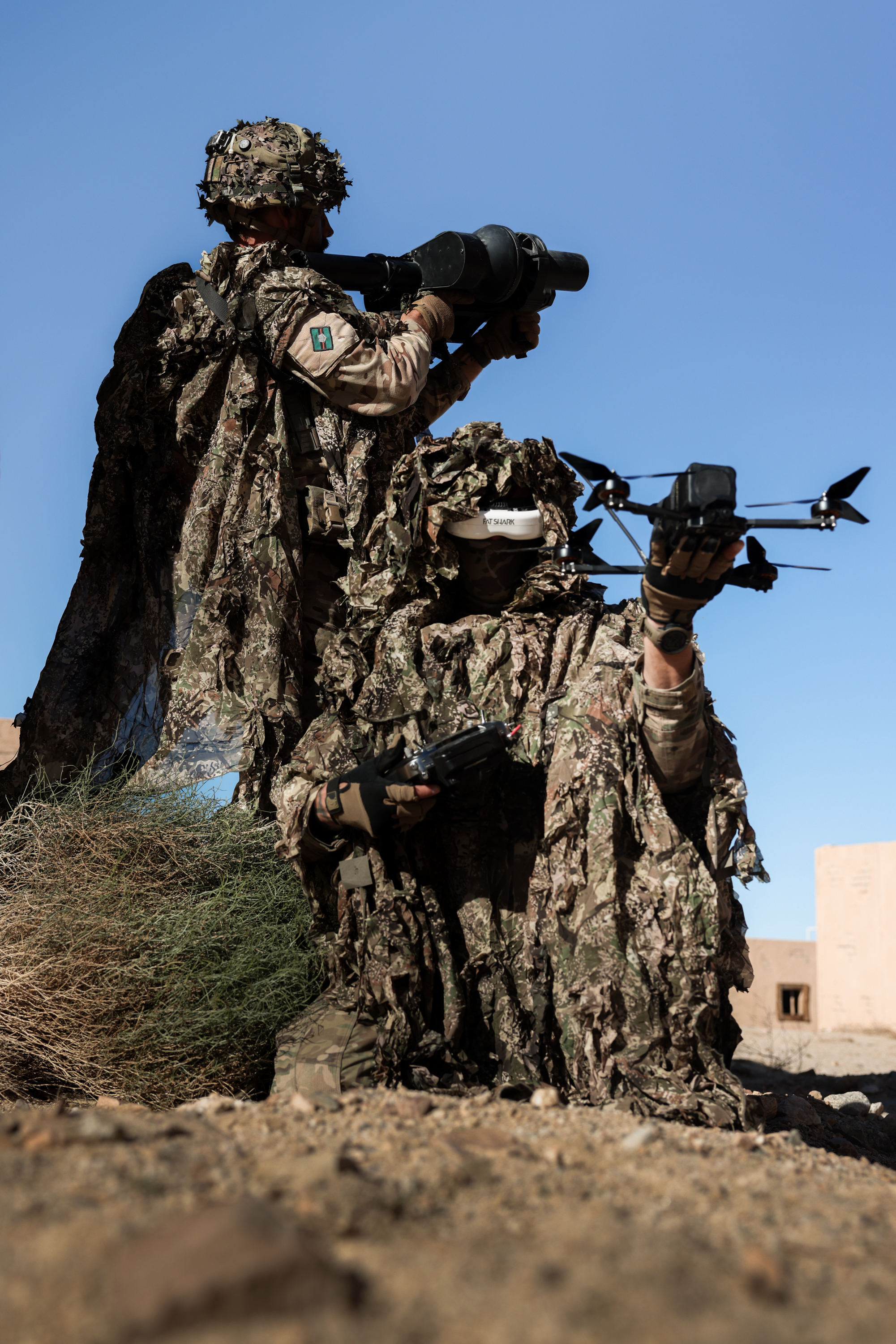 Two soldiers wearing camouflage suits hold a drone and a large gun.