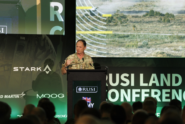 A military soldier is standing at a lectern talking to a room of people.
