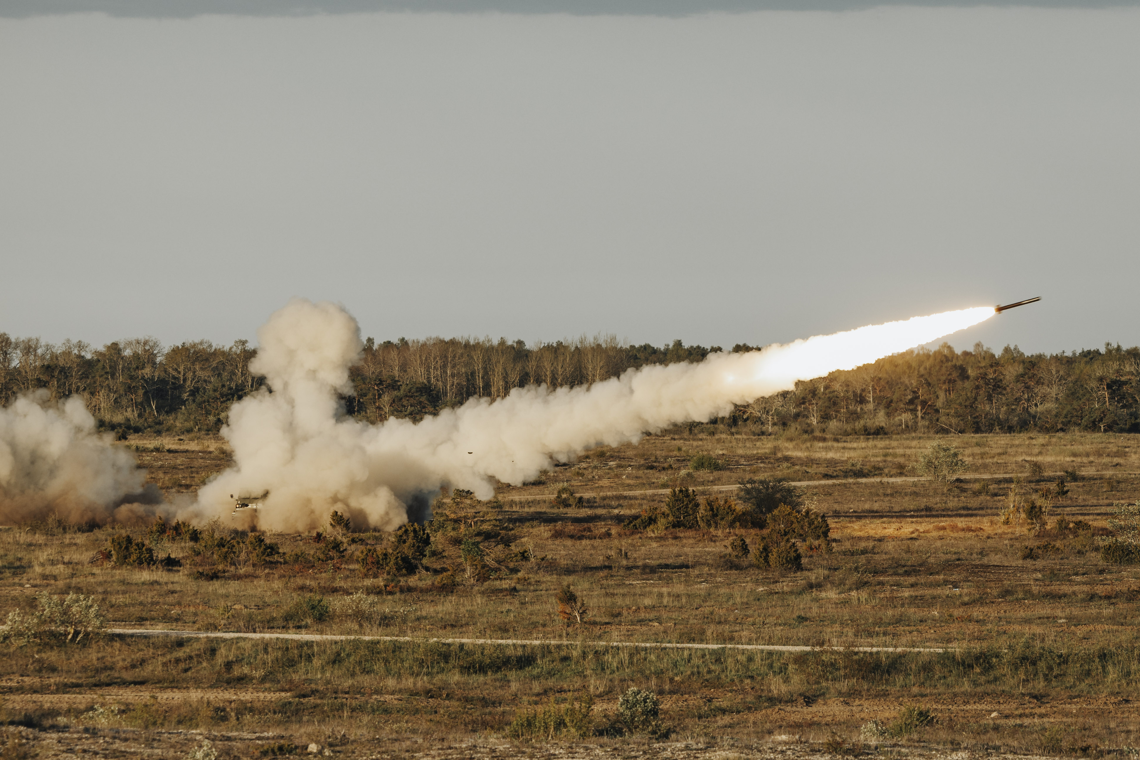 Open and empty tree filled landscape shows a reduced range practise rocket having been fired with a plume of smoke behind it.