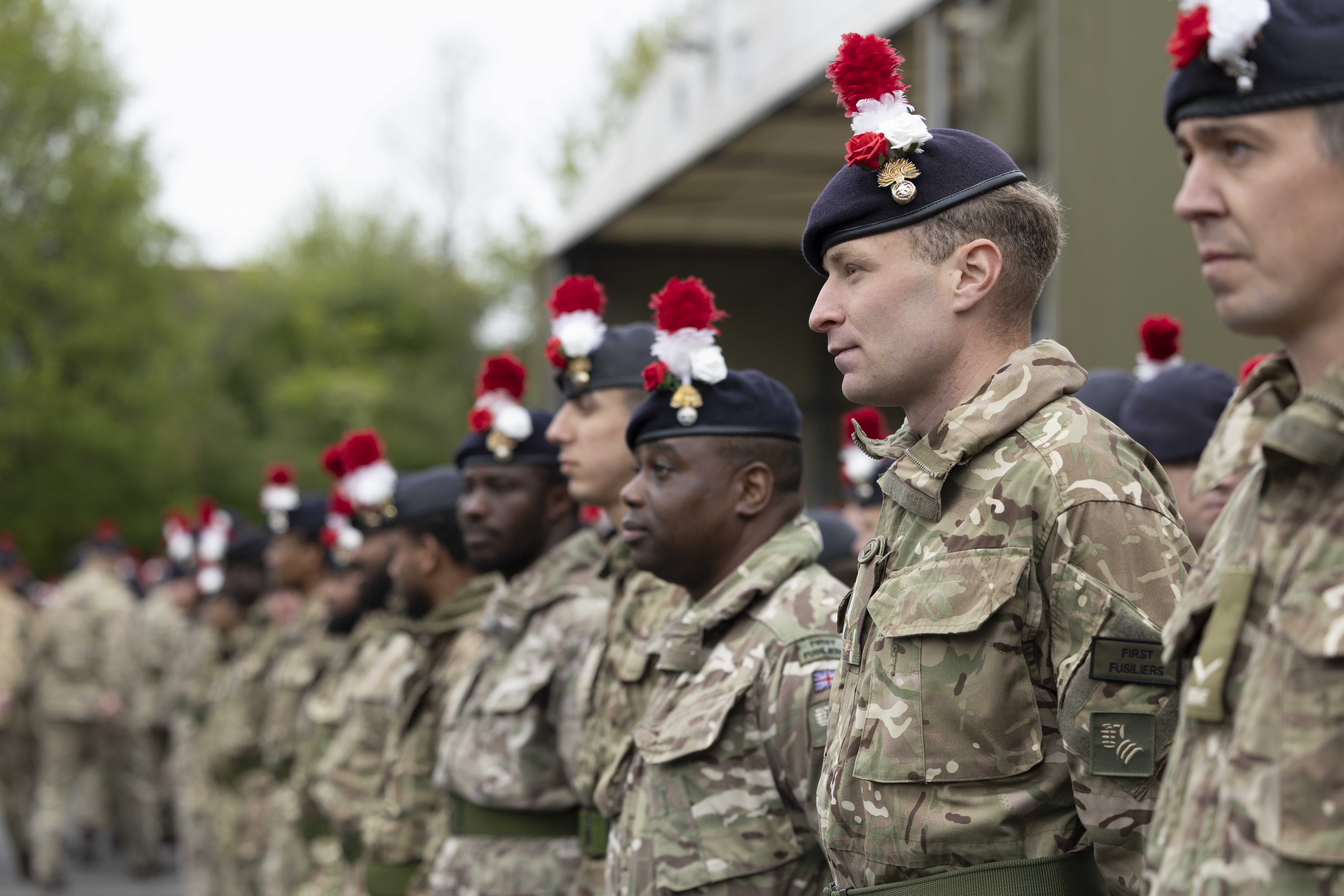Soldiers in camouflage uniforms stand in formation wearing dark berets with red and white plumes outdoors.