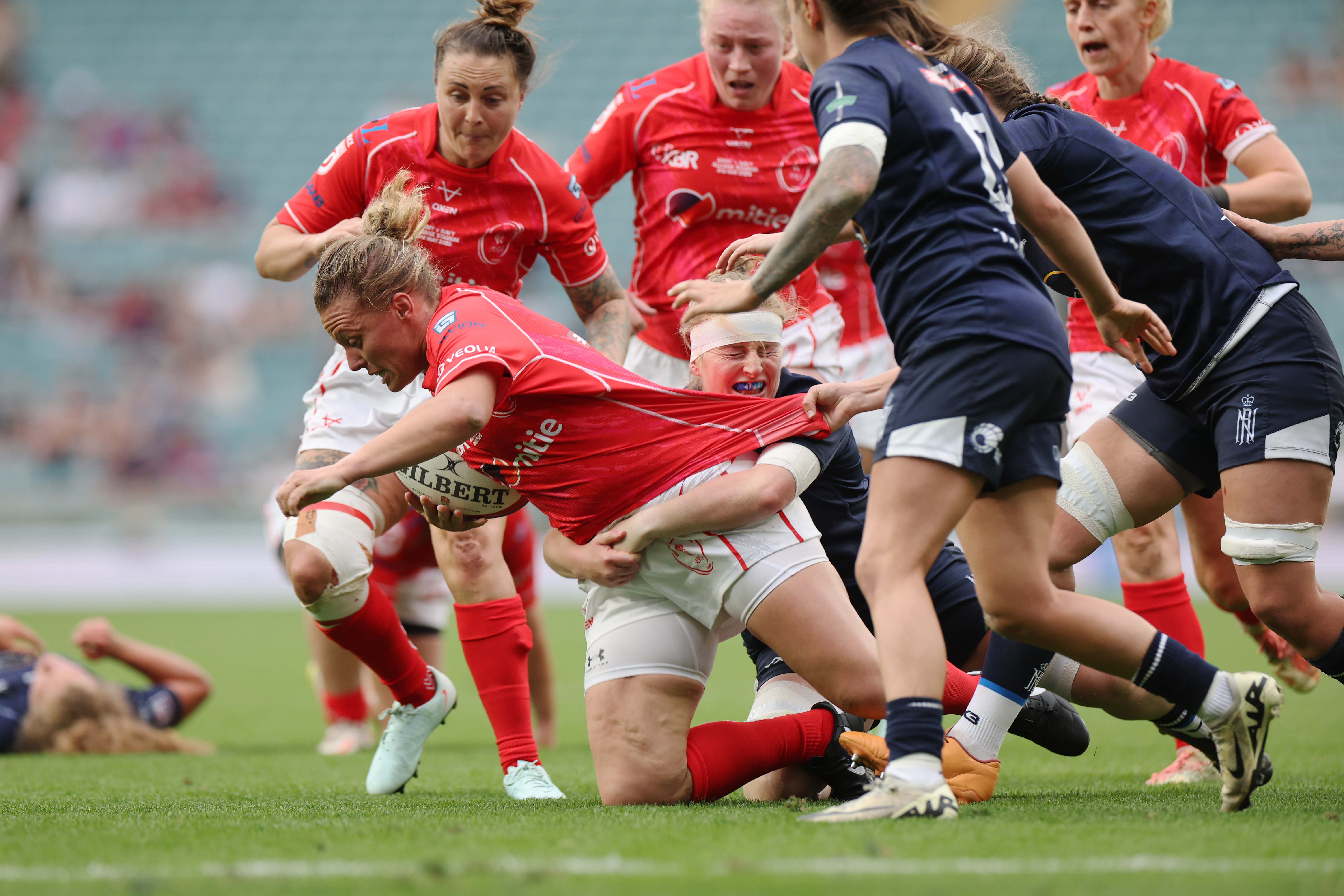 Rugby players in red and navy jerseys engaged in an intense play on a green field. A player in red holds the ball tightly while others tackle and pull their shirt, conveying strong determination and teamwork.
