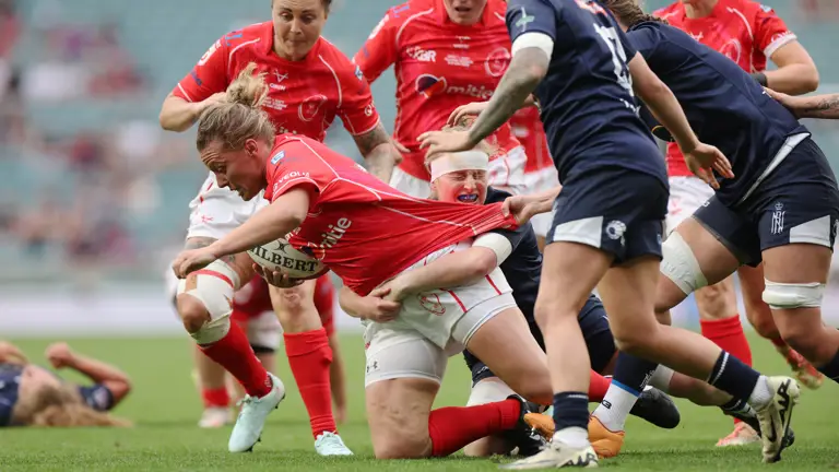 Rugby players in red and navy jerseys engaged in an intense play on a green field. A player in red holds the ball tightly while others tackle and pull their shirt, conveying strong determination and teamwork.