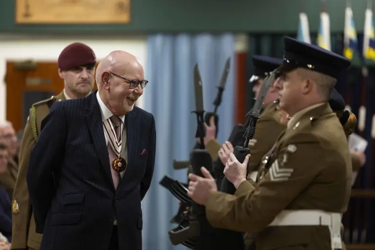 A distinguished elderly man in a striped suit smiles warmly at uniformed soldiers holding rifles with bayonets. Formal setting, atmosphere is respectful and solemn.