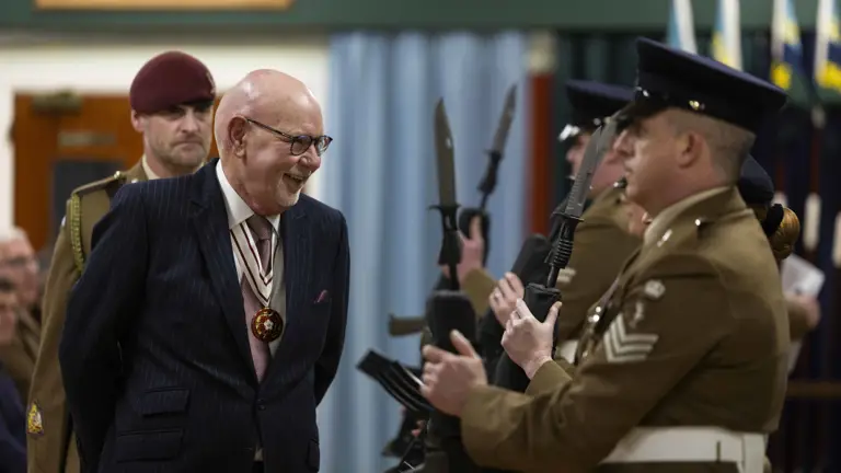 A distinguished elderly man in a striped suit smiles warmly at uniformed soldiers holding rifles with bayonets. Formal setting, atmosphere is respectful and solemn.