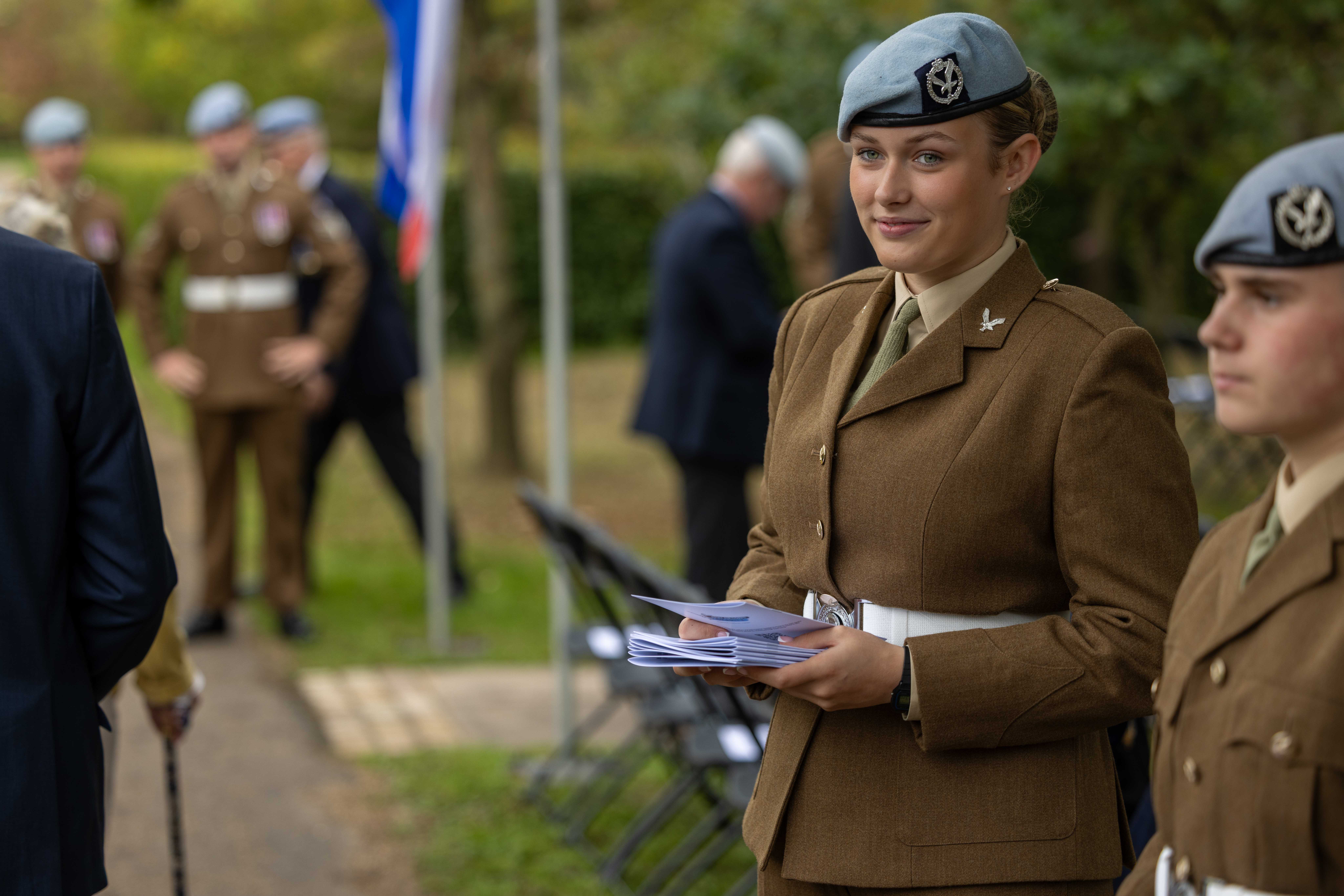 A person in a brown military dress uniform with a light blue beret holds a stack of papers, standing outdoors at a formal event. Other uniformed individuals and a flag are visible in the background.